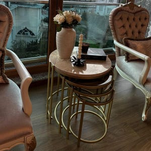 May include: A set of three nesting tables with round tops and gold-toned metal frames. The top table features a light beige marble surface. A white ceramic vase with a bouquet of white roses, a tall bubble candle, and a black book sit on the table.