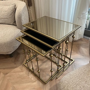 May include: A set of three nesting tables featuring square, reflective mirrored tops and a metallic gold-toned frame. Each table is supported by four slender legs accented with decorative spherical beads at varying heights along the vertical supports.