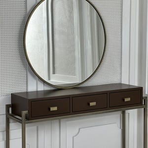 Peut inclure: Un miroir mural rond doté d'un cadre métallique fin, placé au-dessus d'une console rectangulaire brun foncé. La console comporte trois tiroirs horizontaux, chacun équipé d'une petite poignée rectangulaire en métal brossé.