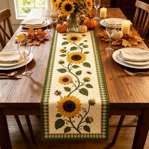 Puede incluir: Un camino de mesa acolchado con un panel central de color crema que presenta un patrón repetitivo de girasoles amarillos, hojas verdes y pequeñas abejas, bordeado por un ribete a cuadros verdes y blancos sobre una mesa de madera.