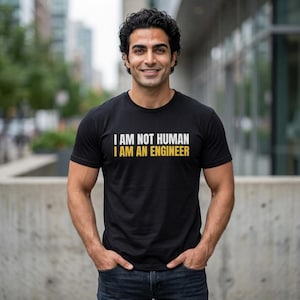Könnte beinhalten: Ein schwarzes Kurzarm-T-Shirt mit dem weißen Text I AM NOT HUMAN in der oberen Zeile und dem gelben Text I AM AN ENGINEER in der unteren Zeile. Das T-Shirt hat einen Rundhalsausschnitt und besteht aus einem einfarbigen schwarzen Stoff.