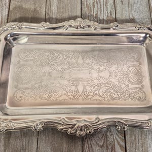 Peut inclure: Un plateau de service rectangulaire de couleur argentée doté d'un bord surélevé orné de volutes et de motifs en forme de coquillages. La surface centrale plate présente un motif floral gravé complexe avec des détails symétriques.