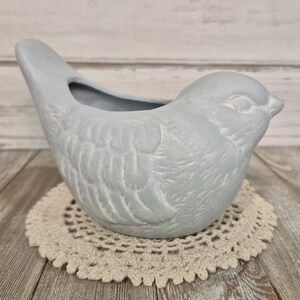 May include: A light grey bird-shaped planter with a matte finish. The ceramic vessel features moulded wing details on the sides, a smooth head with a defined beak, and an open cavity on the back for holding small items or plants.