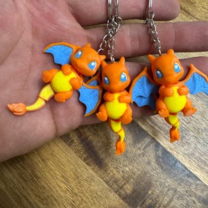 May include: Three orange and yellow dragon-like keychains with blue wings and blue eyes. Each figure features a rounded body, small limbs, and a long tail, attached to a silver-colored metal split ring by a short chain link connector.