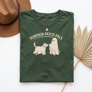 May include: A dark green t-shirt featuring a cream-colored graphic of a dog and a ghost standing together. Above the graphic, the text reads PUMPKIN PATCH PALS in a curved font. A gold-toned necklace rests on the neckline of the shirt.