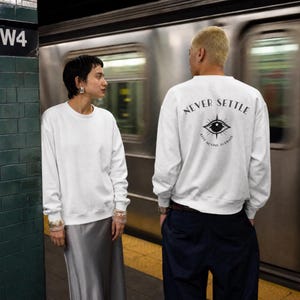 May include: A white long-sleeve crewneck sweatshirt featuring a black graphic on the back. The graphic displays an eye symbol with the text NEVER SETTLE arched above and KEEP MOVING FORWARD curved below. The sweatshirt has a plain white front.