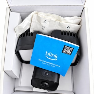 May include: A black Blink wired floodlight camera with two adjustable light panels, packed inside a white cardboard box. Included are white accessory bags labeled A and B, and a blue Blink Wired Floodlight Camera Quick Start Guide booklet.