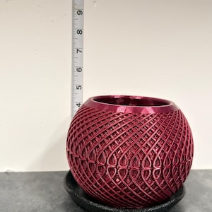 May include: A spherical, metallic magenta planter with a complex, repeating lattice pattern. It sits on a black circular saucer. A vertical measuring tape placed behind the planter indicates a height of approximately 4 inches from the base to the rim.