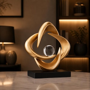 Può includere: Una scultura astratta color oro caratterizzata da forme curve e levigate che si intersecano creando un'apertura centrale. Una sfera di vetro trasparente poggia al centro della struttura dorata, montata su una base rettangolare nera.
