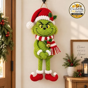 Könnte beinhalten: Ein gehäkelter Wandbehang in Form einer grünen Figur mit schelmischem Gesichtsausdruck, die eine rot-weiße Weihnachtsmütze mit Stechpalme, einen rot-weiß gestreiften Schal mit goldener Glocke und rote, geringelte Schuhe mit weißem Rand trägt.