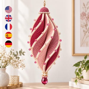May include: A hanging crochet ornament featuring a spiraling, multi-toned pink and cream design. The structure is adorned with small, evenly spaced pink beads along the edges, finished with a cluster of cream pearls and a faceted pink crystal drop.