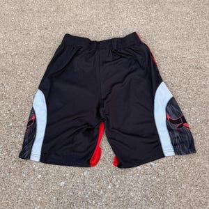 May include: Black athletic shorts featuring a wide elastic waistband, white side panels with a dark gray abstract pattern, and a red bull graphic on each side. The shorts have red accents along the inner leg seams and a smooth, lightweight fabric.