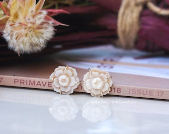 May include: A pair of delicate white flower earrings with gold accents. The earrings are made of carved shell and are displayed on a white surface with a book and flowers in the background.