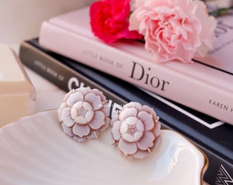 May include: A pair of pink shell flower earrings with silver accents. The earrings are displayed on a white plate with a gold rim.