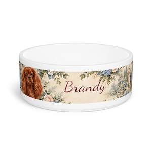 May include: A white ceramic pet bowl featuring a wrap-around design with a brown Cavalier King Charles Spaniel, floral patterns in blue and pink, and the name Brandy written in a dark red script font across the center of the exterior.