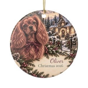 Op de afbeelding: Een ronde keramische kerstornament met een bruine Cavalier King Charles Spaniël met een donkerrode strik, tegen een besneeuwd huisje met dennentakken en bessen. De tekst luidt Oliver en Christmas 2026 in donkere letters.