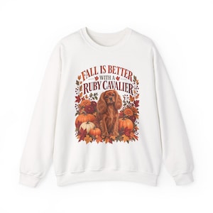 May include: A white crewneck sweatshirt featuring a graphic print of a brown Cavalier King Charles Spaniel surrounded by orange pumpkins, red and yellow autumn leaves, and flowers, with the text "FALL IS BETTER WITH A RUBY CAVALIER" above.