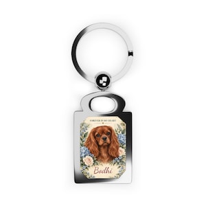 May include: A silver-toned metal keychain featuring a rectangular pendant with a rounded top. The pendant displays a portrait of a brown Cavalier King Charles Spaniel surrounded by blue and white flowers, with the text "FOREVER IN MY HEART" and "Bodhi" printed below.