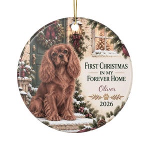 Puede incluir: Un adorno redondo de cerámica con un Cavalier King Charles Spaniel marrón sentado en un porche. El texto dice First Christmas in my Forever Home, Oliver, y 2026. El fondo muestra una puerta decorada, ramas de pino y nieve.