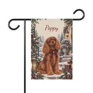 May include: A rectangular garden flag featuring a brown Cavalier King Charles Spaniel wearing a red plaid bow with a bell. The scene includes snowy evergreen branches, a lantern, and a gift box. The name Poppy is printed in red script at the top.