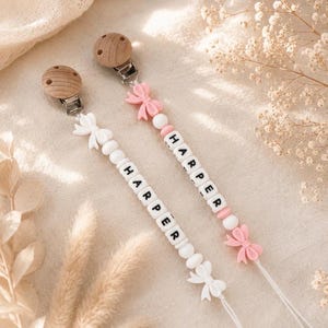 May include: Two personalised dummy clips featuring round wooden fasteners with metal clips. One clip has white beads and a white bow, the other has pink beads and a pink bow. Both feature black letter beads spelling the name HARPER in a vertical sequence.