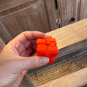 May include: A bright orange fidget cube puzzle toy composed of multiple interconnected square segments with bevelled edges. The object features a matte finish and a geometric design that allows the individual parts to fold and shift into various shapes.