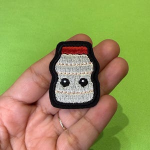 May include: An embroidered patch shaped like a small bottle with a red cap and a silver body. The bottle features two small black circular eyes and horizontal stitching details. The entire design is outlined with a thick black border.