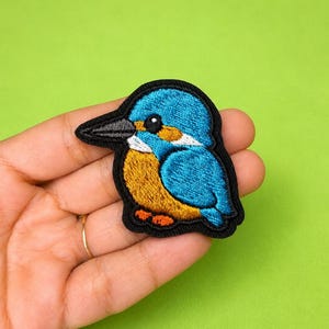 Op de afbeelding: Een geborduurde patch met een gestileerde ijsvogel met felblauwe veren, een goudoranje borst, een witte keelvlek en een donkergrijze snavel. De vogel is omlijnd met zwart garen en heeft een klein zwart oog met een witte reflectie.