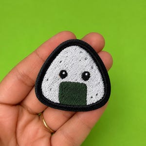 May include: An embroidered patch shaped like a triangular onigiri rice ball. The patch features a white rice texture with two black circular eyes and a dark green rectangular nori seaweed strip at the base, all outlined with a thick black border.