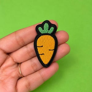 May include: An embroidered iron-on patch shaped like a carrot. The carrot has a bright orange body with thin black horizontal lines for texture, topped with three green leaves. The entire design is outlined with a thick black border.