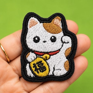 Könnte beinhalten: Ein gestickter Aufnäher mit einer weißen Katze mit braunen Flecken, einem roten Halsband mit goldener Glocke und einer gelben Münze mit schwarzem japanischen Text. Die Katze hat eine erhobene Pfote und einen dicken, schwarzen Rand.