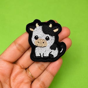 Può includere: Una toppa ricamata raffigurante una mucca dei cartoni animati con corpo a macchie bianche e nere, muso color cuoio e piccole corna. Il disegno è delineato da uno spesso filo nero, creando la forma di un vitello seduto con grandi occhi scuri.