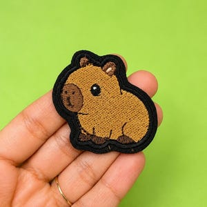 May include: An embroidered patch featuring a cartoon capybara with light brown fur, a darker brown snout, and small ears. The capybara has a black eye and is outlined with a thick black border, creating a distinct shape against the background.