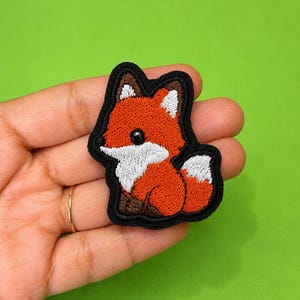 Op de afbeelding: Een geborduurde patch met een gestileerde vos met oranje vacht, een witte borst, witte oortoppen en een staart met een witte punt. De vos heeft een kleine zwarte neus en een zwart oog, alles omlijnd met een dikke zwarte rand op een stoffen ondergrond.