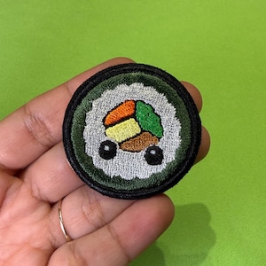 Pode incluir: Um emblema bordado circular com um design de rolo de sushi. O emblema tem uma borda preta, um centro de arroz branco e segmentos bordados coloridos em laranja, amarelo, castanho e verde, com dois pequenos círculos pretos como olhos.