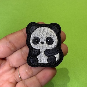 Könnte beinhalten: Ein gestickter Aufnäher in Form eines stilisierten Pandas. Der Panda hat ein weißes Gesicht und einen weißen Körper mit schwarzen Ohren, schwarzen Augenpartien und schwarzen Gliedmaßen. Das Design ist von einem dicken schwarzen Rand umgeben.