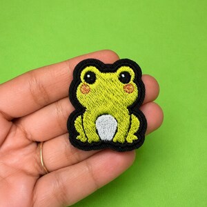 Può includere: Una toppa ricamata raffigurante una rana in stile cartoon con pelle verde brillante, grandi occhi neri, piccole guance rosa e una pancia bianca. La rana è delineata da un bordo nero spesso, che crea una forma distinta e ben definita.