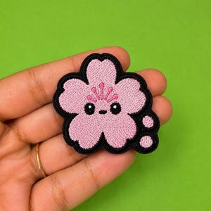 Op de afbeelding: Een roze geborduurde patch in de vorm van een kersenbloesem met vijf bloemblaadjes en een zwarte rand. De bloem heeft een schattig gezichtje met twee zwarte ronde ogen, een klein horizontaal streepje als mond en drie roze meeldraden.