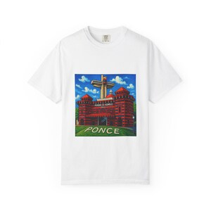 Puede incluir: Una camiseta blanca de manga corta con un estampado gráfico del Parque de Bombas en Ponce. La ilustración muestra el edificio de rayas rojas y negras con una gran cruz, cielo azul y la palabra PONCE impresa en blanco sobre el césped verde.