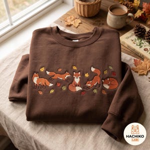 Puede incluir: Una sudadera de cuello redondo en color marrón oscuro con un diseño bordado de cinco zorros naranjas y blancos en varias posturas, intercalados con pequeñas hojas de otoño de colores dispersas por la zona del pecho de la prenda.