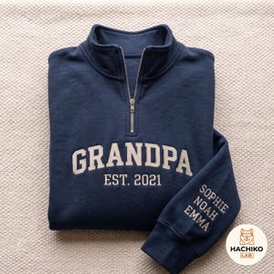 Puede incluir: Una sudadera azul marino con cremallera de un cuarto y el texto bordado GRANDPA EST. 2021 en blanco en el pecho. La manga está bordada con los nombres SOPHIE, NOAH y EMMA en blanco. La prenda cuenta con un cierre de cremallera de metal plateado.