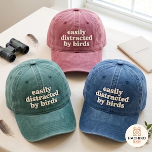 Op de afbeelding: Drie baseballpetten in oudroze, blauwgroen en blauw, elk met de geborduurde witte tekst "easily distracted by birds" op de voorkant. De petten hebben een vintage gewassen afwerking, een gebogen klep en een knoopje bovenop de kroon.