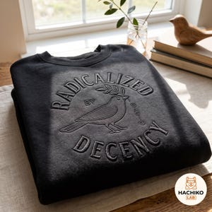 May include: A black crewneck sweatshirt featuring an embossed design on the chest. The graphic includes a bird holding an olive branch, surrounded by the text "RADICALIZED BY BASIC DECENCY" in a circular arrangement with a clean, raised texture.