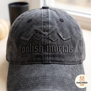 Op de afbeelding: Een antracietgrijze baseballpet met een verwassen, verweerde afwerking. De voorkant is voorzien van donkergrijs borduurwerk van vier vliegende vleermuizen boven de tekst "foolish mortals" in een gotisch lettertype op het voorpaneel.