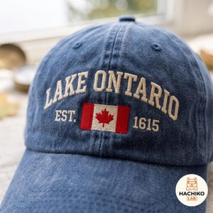 Könnte beinhalten: Eine blaue Baseballkappe aus Denim mit dem gestickten Text LAKE ONTARIO EST. 1615, zentriert über einer kleinen kanadischen Flagge mit einem roten Ahornblatt. Die Kappe hat eine strukturierte Krone mit gestickten Ösen und einen gebogenen Schirm.