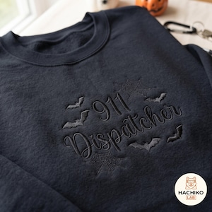 Peut inclure: Un sweat-shirt noir à col rond avec une broderie gris foncé du texte 911 Dispatcher en police cursive, entouré de motifs de toiles d'araignée et de chauves-souris. Le motif est centré sur la poitrine du vêtement à manches longues.