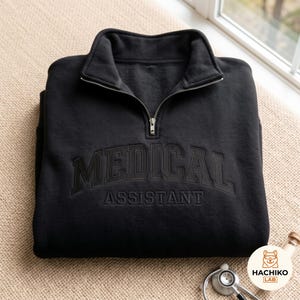 Puede incluir: Una sudadera negra con cremallera de cuarto de cuello y el texto MEDICAL ASSISTANT bordado en hilo negro a juego en el pecho. La prenda cuenta con un cuello alto con cierre de cremallera de metal plateado y mangas largas, presentada de forma plegada.