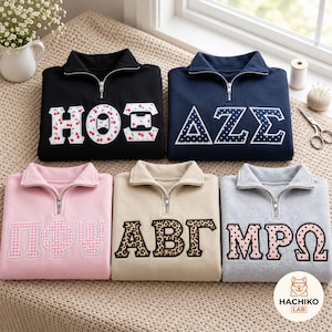 May include: Five quarter-zip sweatshirts in black, navy, pink, beige, and grey. Each features large Greek letter appliqués with various patterns including cherries, white polka dots, pink gingham, leopard print, and black polka dots on a light background.