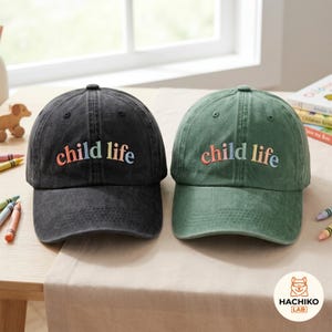 Puede incluir: Dos gorras de béisbol, una en negro carbón y otra en verde apagado, ambas con el texto "child life" bordado en una fuente arqueada multicolor en los paneles frontales. Cada gorra tiene una corona estructurada y una visera curva.