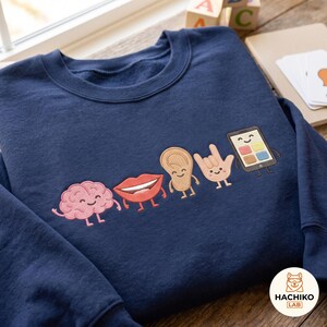 May include: A navy blue sweatshirt featuring an embroidered graphic of five cartoon characters: a pink brain, red lips, a tan ear, a tan hand making a sign language gesture, and a black smartphone with a colourful screen, all with smiling faces.